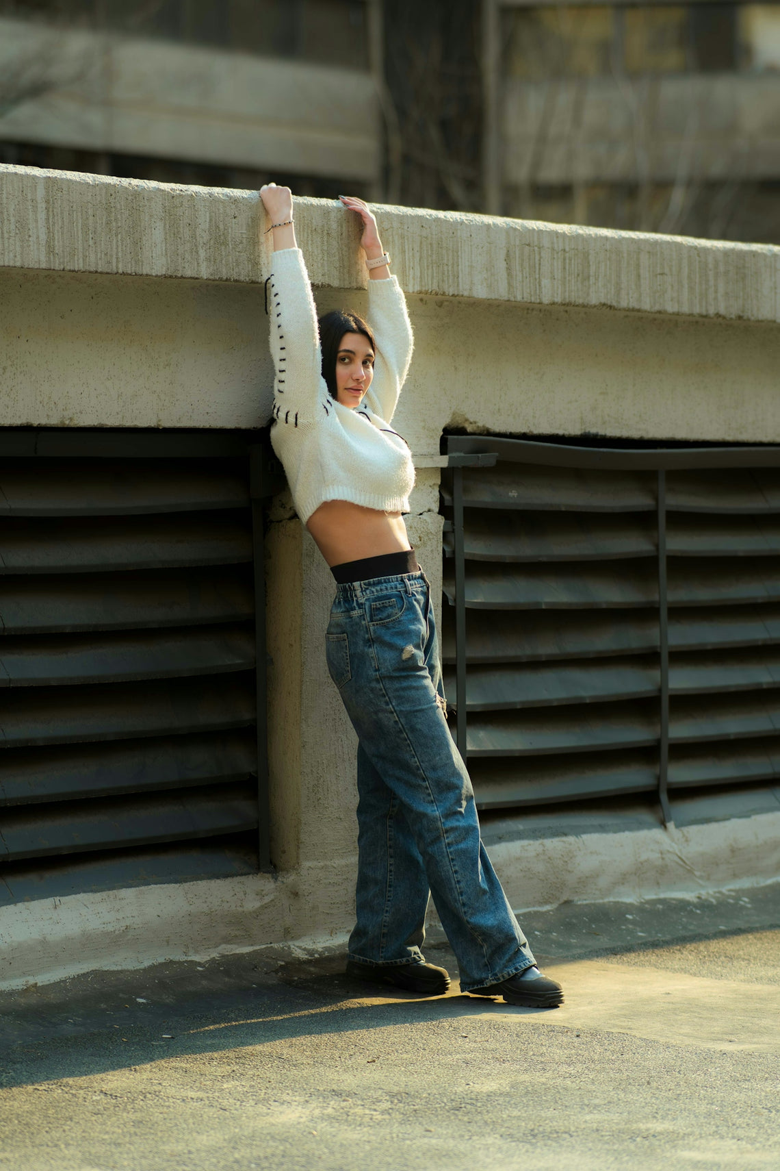Young woman leaning against concrete wall, wearing jeans and sweater.