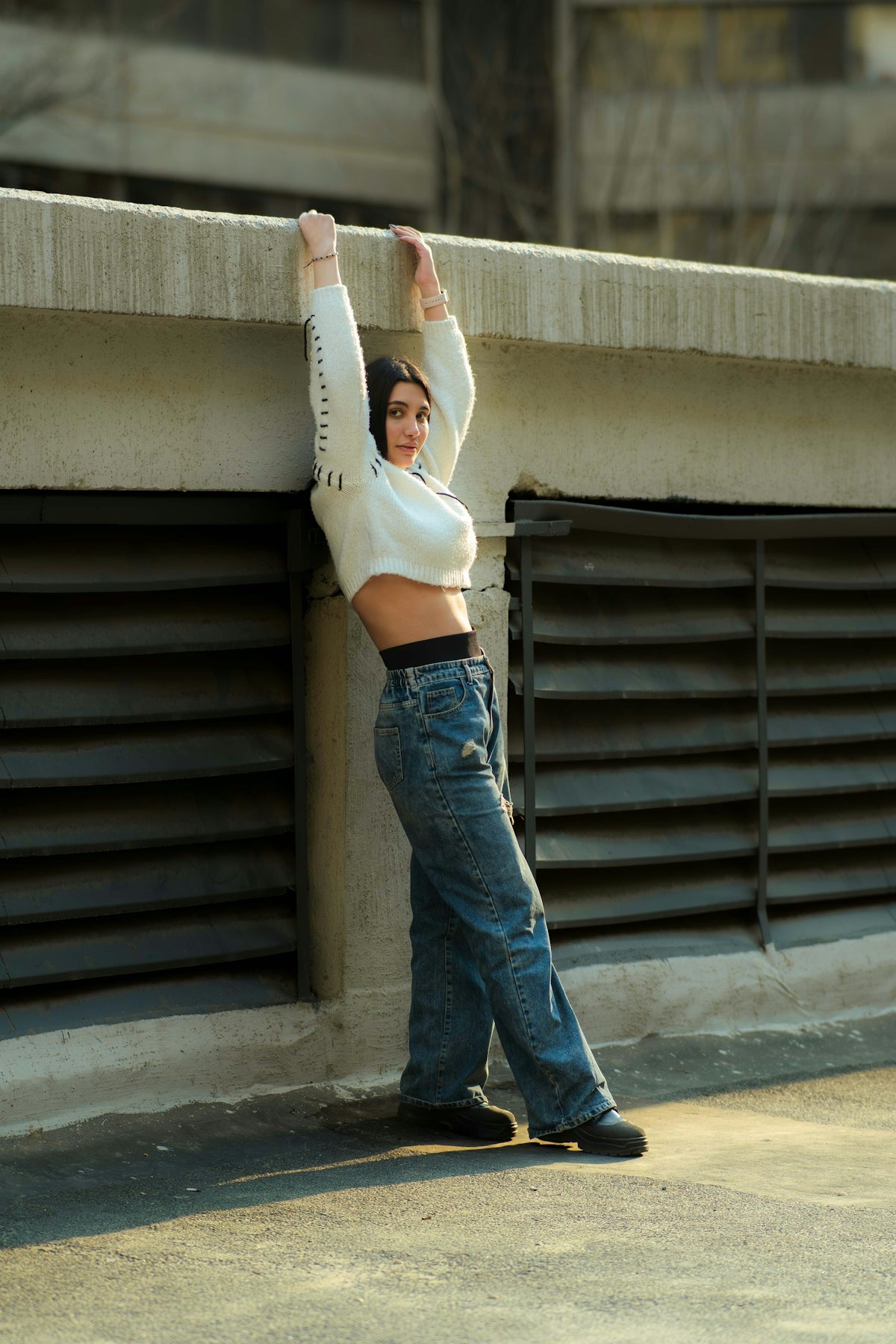 Young woman leaning against concrete wall, wearing jeans and sweater.