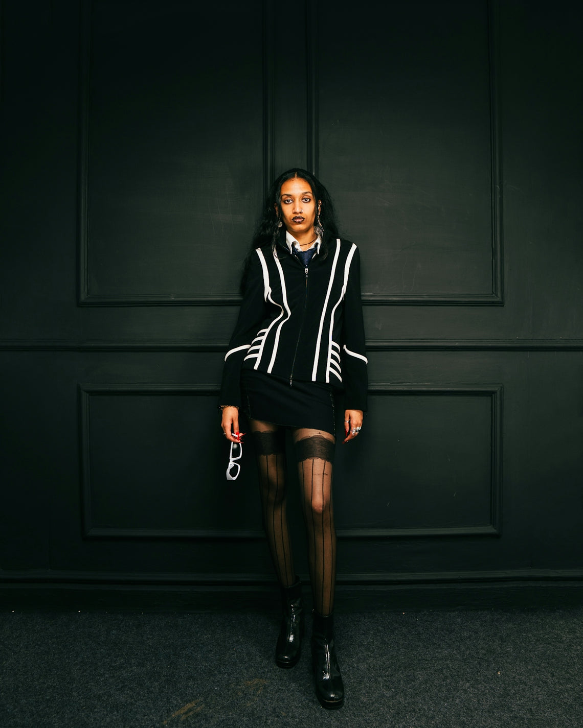A woman in stylish clothing stands against a dark wall.