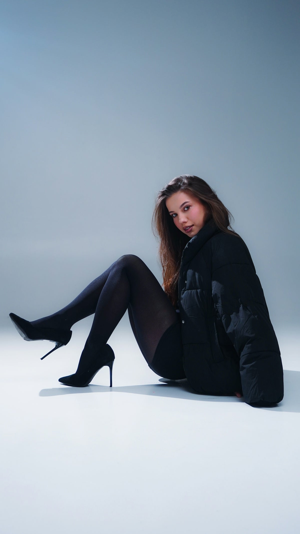 A woman in a black puffer jacket and heels sits.