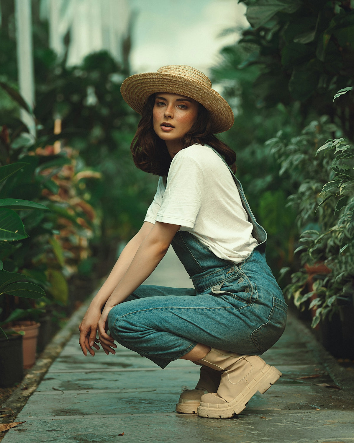 Woman in straw hat and overalls crouching among green plants.